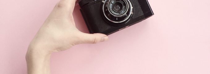 an arm is reaching upwards with the hand holding a black camera against a pink background.